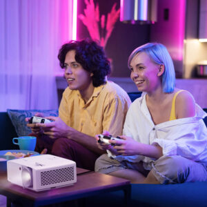 Young man and women playing video games sitting on a couch, with a projector on the coffee table