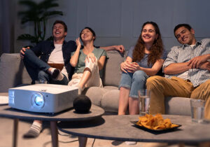 Four young adults looking at a screen, with a projector placed on the coffee table.