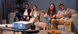 Four young friends sitting on a couch watching a movie and laughing, with a projector on the coffee table.