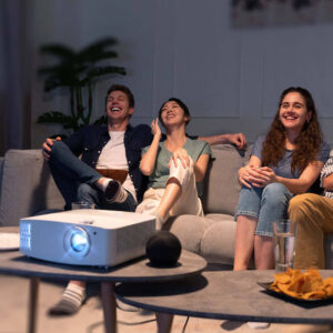 Four young friends sitting on a couch watching a movie and laughing, with a projector on the coffee table.