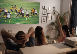 A man and two women sitting on a couch watching a football game being projected on the wall.