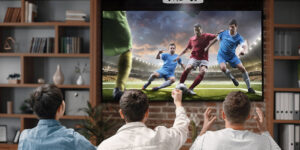 Three men in a living room with a projector mounted at the ceiling, displaying a soccer game.