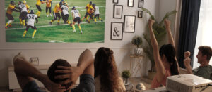 Four friends sitting on the couch watching football on a projector screen. The projector is behind them on a table.